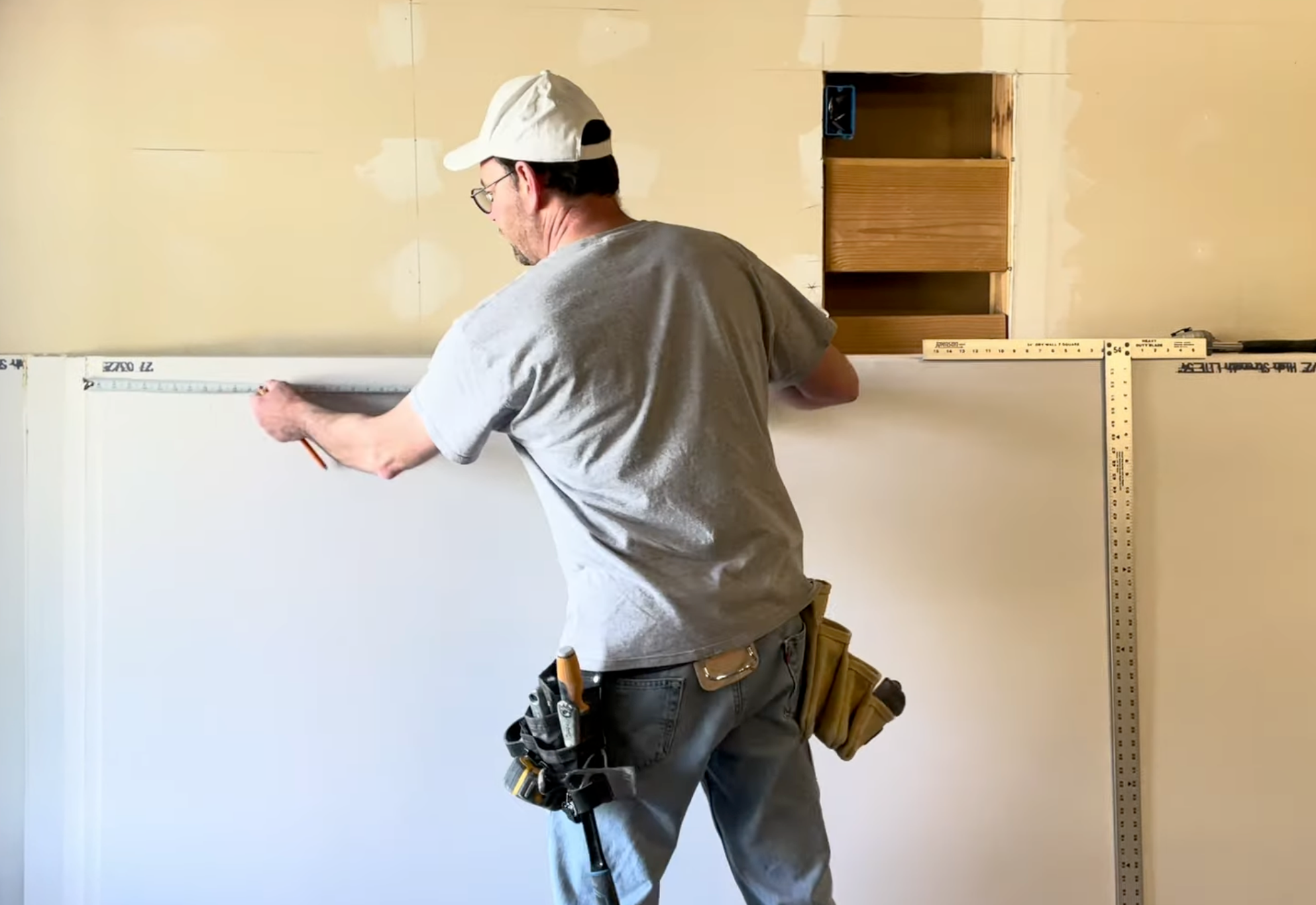 A man wearing a white cap, gray t-shirt, and jeans is shown from behind, measuring a white drywall panel with a metal tape measure in an unfinished room with beige drywall and an exposed wood stud.