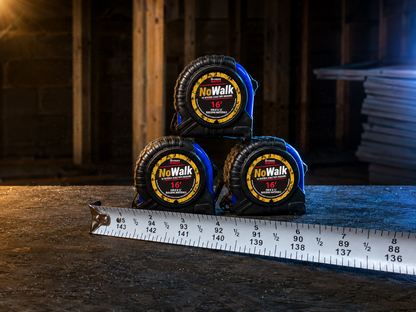 Three NoWalk 16-inch tape measures stacked in a pyramid on a wooden workbench, with a metal measuring tape ruler laid out in front, set against a dark workshop background.